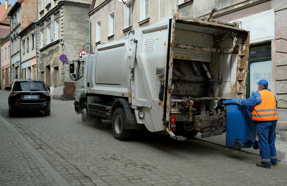 A worker wearing a red and yellow uniform is operating a large, red waste collection vehicle on a street. The rear of the vehicle features various warning and identification decals, along with a refle