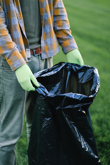 A person wearing a yellow and gray plaid jacket, gray trousers, and bright green gloves is holding an open black plastic rubbish bag outdoors. The bag is partially filled with waste and positioned ove