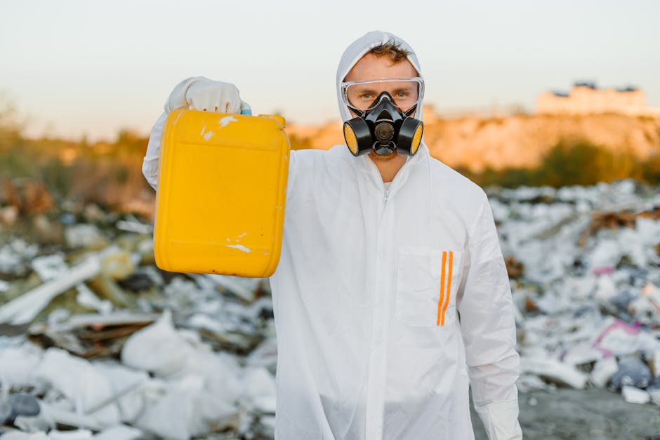 A person wearing a white hazmat suit with orange stripes, protective goggles, and a black respirator mask stands in an outdoor landfill site or rubbish dump. They are holding a yellow plastic containe