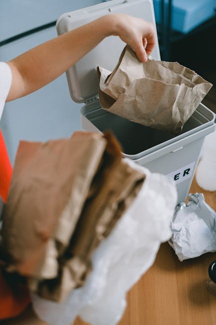 A person's hand is seen placing a brown paper bag filled with waste into an open, white rubbish bin that has a lid positioned nearby. The bin appears to be made of plastic and is situated on a light-c
