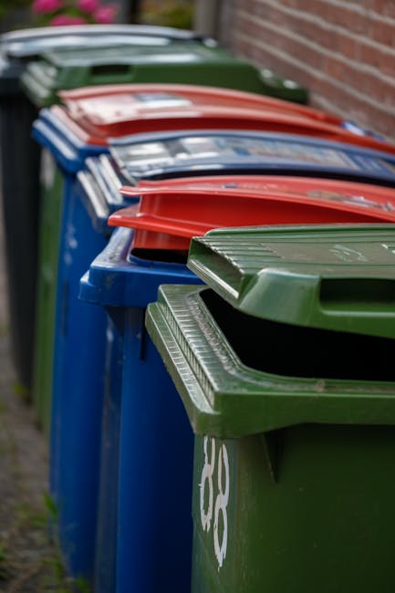 A person's hand is seen placing a brown paper bag filled with waste into an open, white rubbish bin that has a lid positioned nearby. The bin appears to be made of plastic and is situated on a light-c