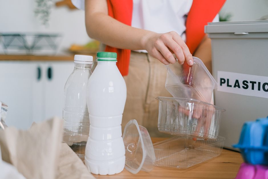 A person wearing a white shirt and an orange vest is placing a clear plastic food container into a recycling bin labeled 'PLASTIC' in a domestic setting. On the wooden table in front of them are two e